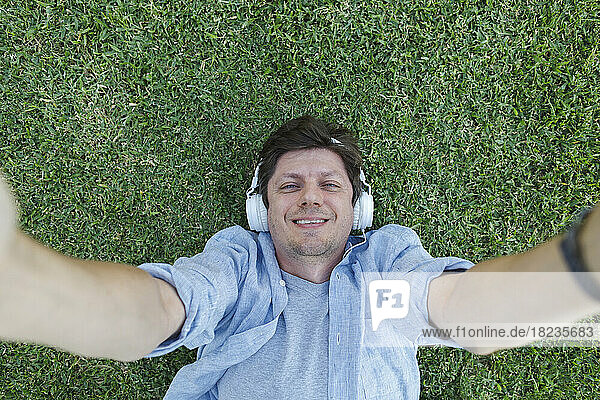 Glücklicher Mann mit Kopfhörern  der ein Selfie macht und im Gras liegt