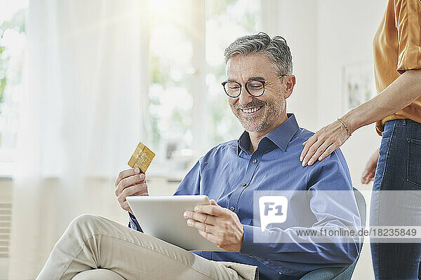 Happy man holding credit card doing online shopping over tablet PC with hand of woman on shoulder at home