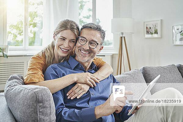 Happy woman embracing man holding tablet PC from behind on sofa at home