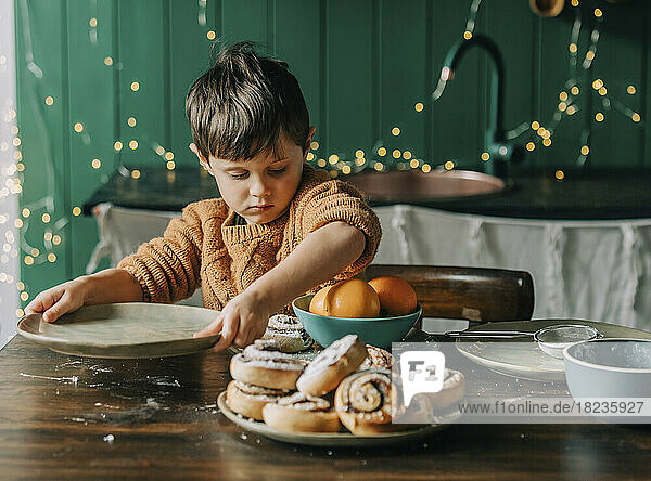 Boy holding plate by cinnamon buns and oranges in bowl at home