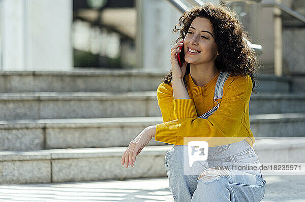 Smiling young woman with curly hair talking over mobile phone on staircase