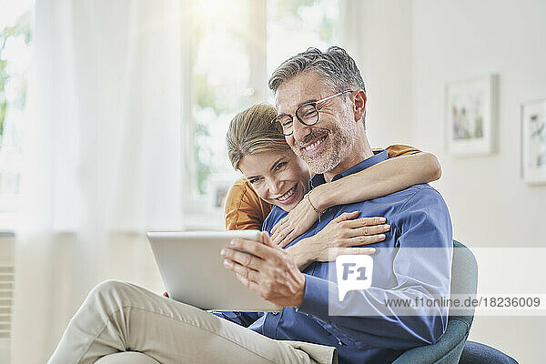 Glücklicher Mann zeigt Frau im Sessel zu Hause Tablet-PC