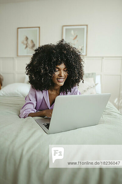 Smiling woman using laptop lying on bed in bedroom