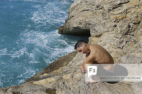 Shirtless man hugging knees on rock near sea