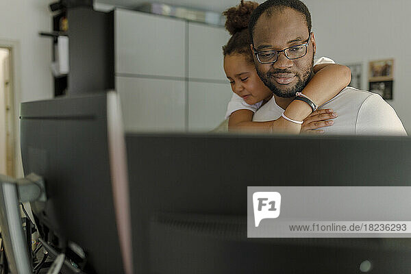 Smiling freelancer working on computer with daughter hugging him from behind at home