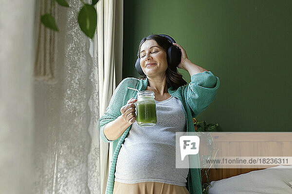 Smiling pregnant woman listening music through wireless headphones holding green smoothie at home