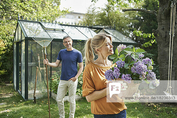 Lächelnde Frau duftet nach Blumen auf einer Topfpflanze  im Hintergrund steht ein Mann im Garten