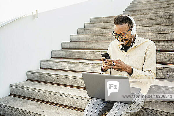 Happy freelancer text messaging through smart phone sitting with laptop on steps