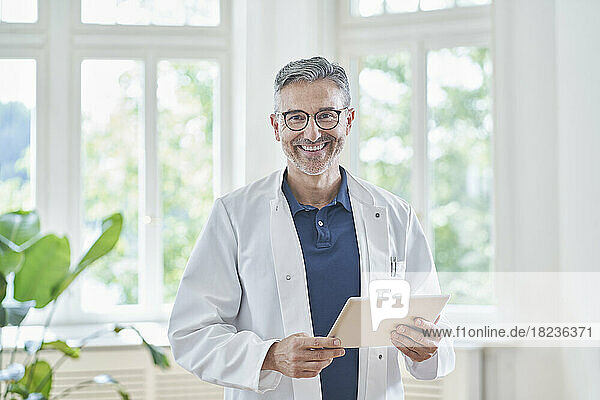 Happy mature doctor standing with tablet PC in medical practice