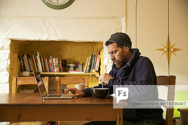 Freelancer having meal using laptop at desk in home