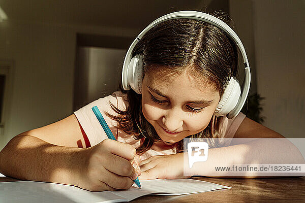 Smiling girl wearing wireless headphones writing in book at desk
