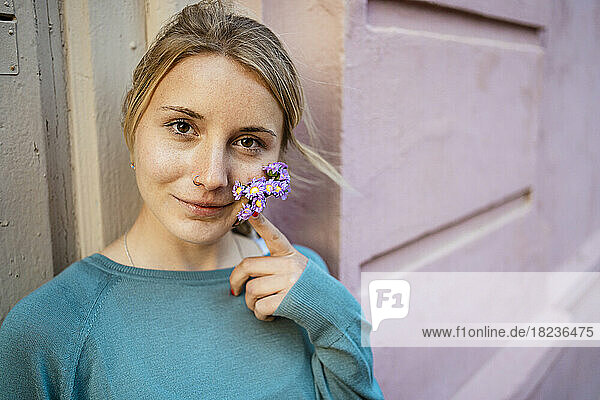 Smiling young woman holding small flowers on face in front of wall