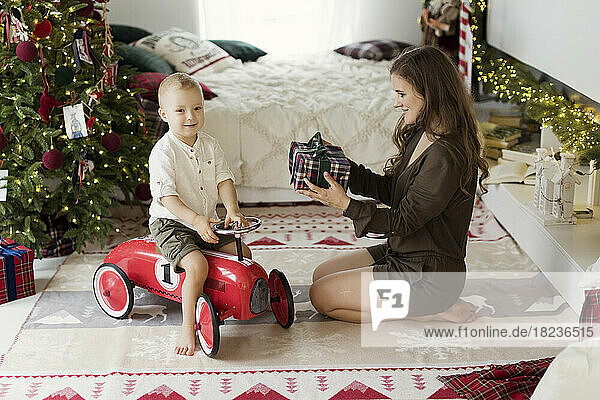Smiling woman giving gift to son sitting on toy car at home