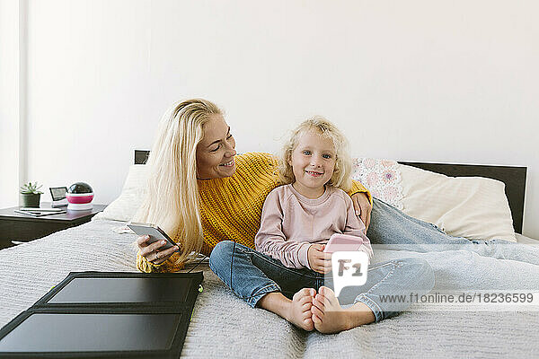 Lächelnde Mutter und Tochter sitzen mit Smartphones auf dem Bett im Schlafzimmer