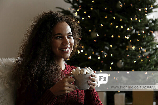 Smiling young woman with cup of marshmallow cocoa sitting in front of Christmas tree
