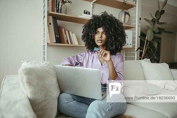 Nachdenkliche Frau mit Laptop und Kreditkarte sitzt im Wohnzimmer