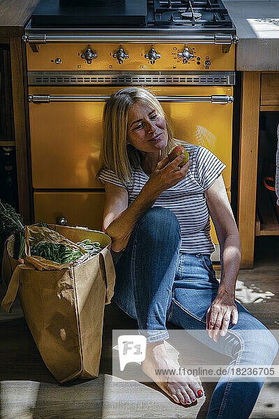 Mature woman eating apple sitting in front of stove at home