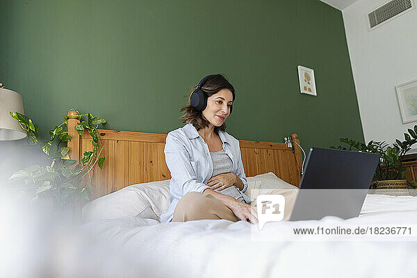 Smiling pregnant businesswoman wearing wireless headphones using laptop on bed in bedroom