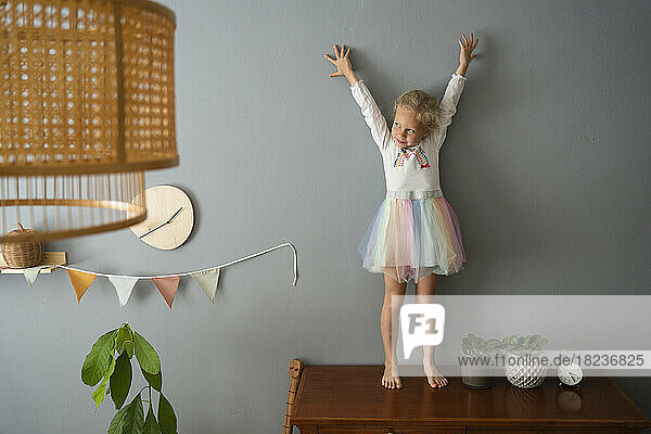 Cute girl standing with hands raised on table at home