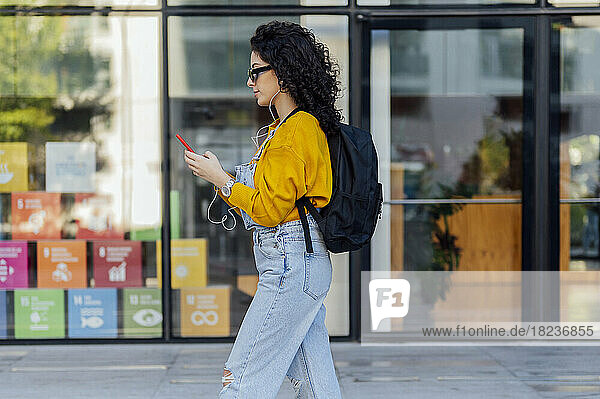Woman listening to music through in-ear headphones and using smart phone in front of glass building