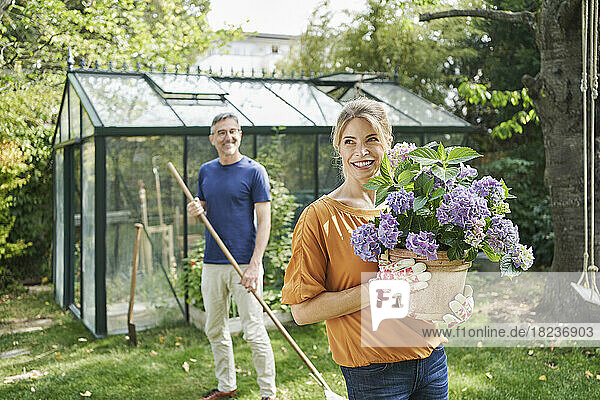 Happy woman holding flowering potted plant with man in background at garden