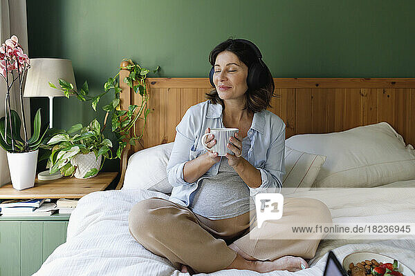 Lächelnde Frau hört Musik über kabellose Kopfhörer und trinkt Tee auf dem Bett im Schlafzimmer