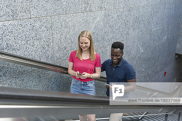 Smiling man and woman using smart phones moving up on escalator by wall