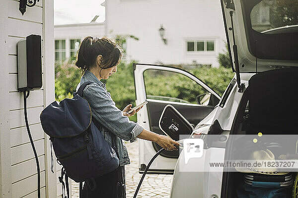 Seitenansicht einer Frau  die beim Aufladen eines Elektroautos ihr Smartphone benutzt
