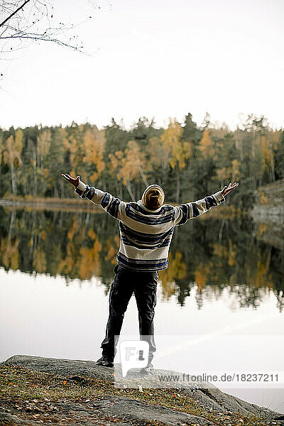 Rear view of carefree man with arms outstretched standing on rock against lake