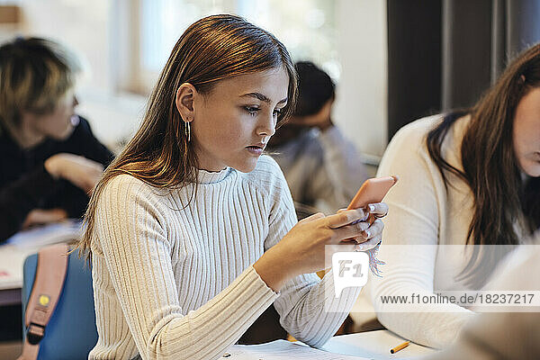 Teenage girl with brown hair using smart phone sitting by female friend at desk in classroom