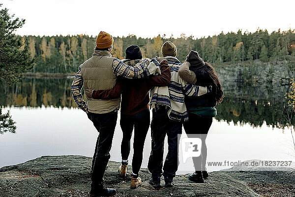 Rear view of male and female friends standing with arm around while looking at lake