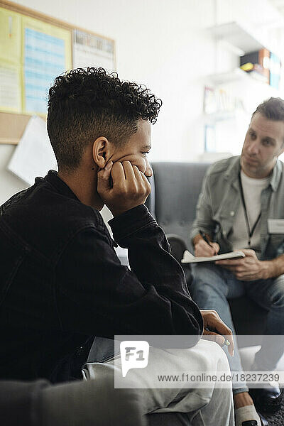 Depressed boy sitting with head in hand by male counselor discussing at school office