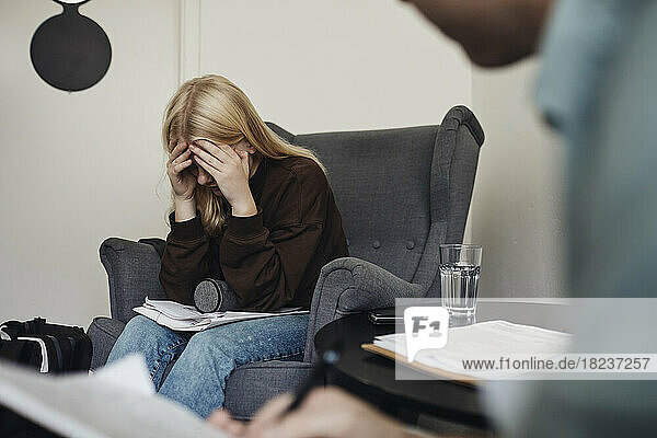 Blond teenage girl with head in hands sitting on chair while discussing with male counselor at school office