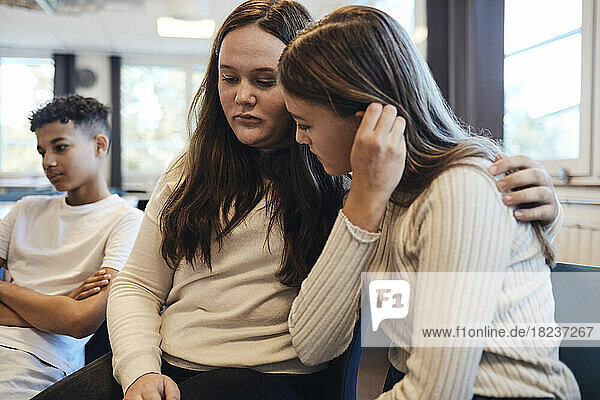 Sad teenage girl sitting with arm around female friend while consoling her in group therapy