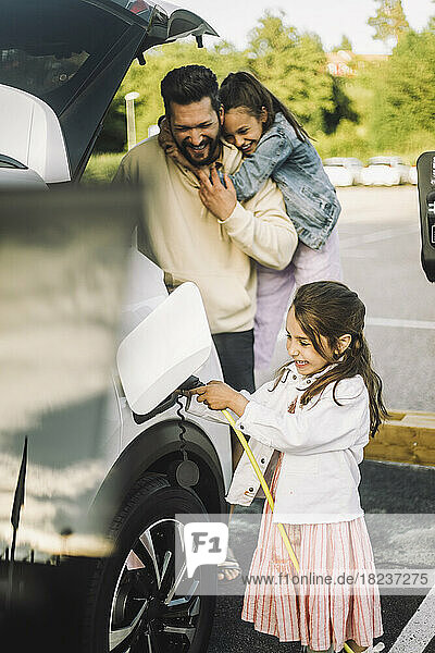 Glücklicher Vater mit Töchtern  die ihr Elektroauto an einer Tankstelle aufladen