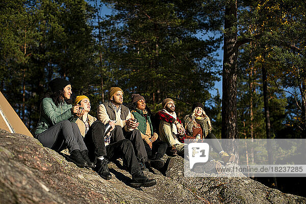 Männliche und weibliche Freunde genießen das Sonnenlicht  während sie auf einem Felsen im Wald sitzen