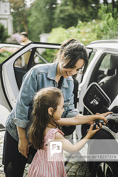 Lächelnde Mutter  die ihrer Tochter beim Aufladen eines Elektroautos hilft