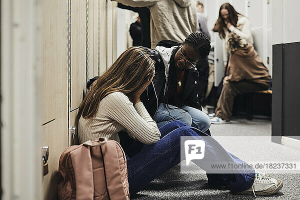 Teenager-Mädchen im Gespräch mit einer depressiven Freundin  die in einem Schulkorridor neben einem Rucksack sitzt