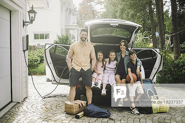Porträt einer glücklichen Familie  die im Kofferraum eines Elektroautos vor dem Haus sitzt