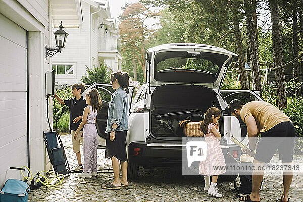 Familie beim Aufladen des Gepäcks im Kofferraum eines Elektroautos vor dem Haus