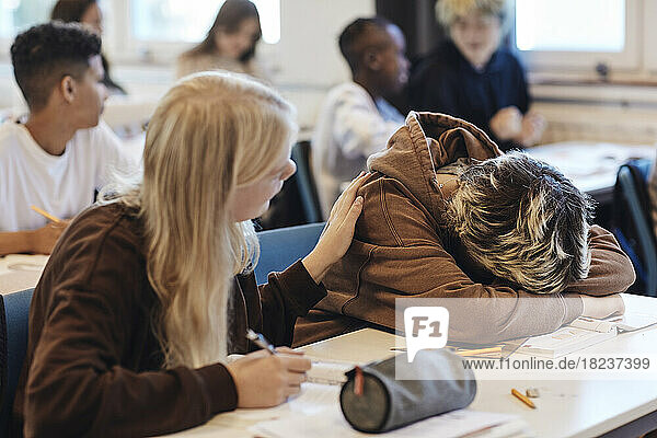 Blondes Teenager-Mädchen tröstet eine deprimierte Freundin  die ihren Kopf auf den Schreibtisch im Klassenzimmer legt