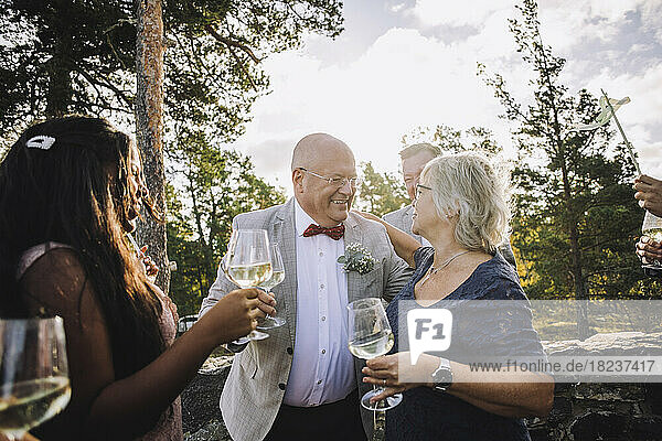 Lächelnde ältere Frau und Bräutigam mit Weingläsern  die sich bei der Hochzeit umarmen