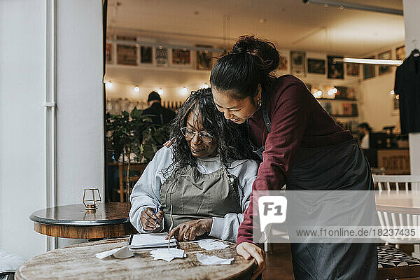 Lächelnde junge Besitzerin  die neben einem Kollegen steht und Tagebuch liest  während sie am Tisch in einem Café sitzt