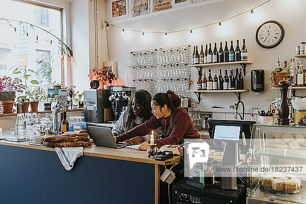 Lächelnde junge Besitzerin  die mit einem Kollegen an der Kasse eines Cafés über einen Laptop diskutiert