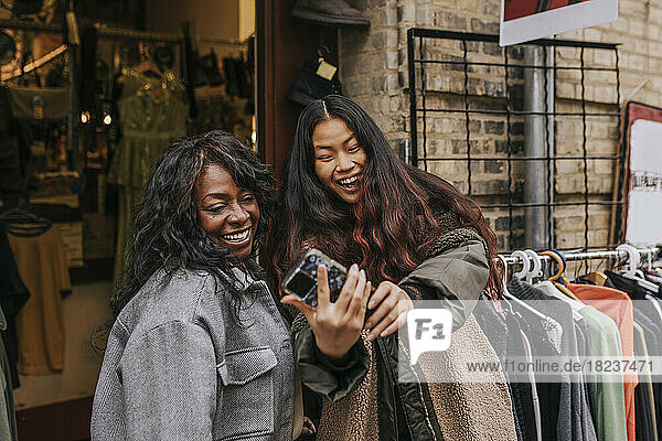 Glückliche Freundinnen  die ein Selfie mit ihrem Smartphone machen  während sie sich vor einem Bekleidungsgeschäft vergnügen
