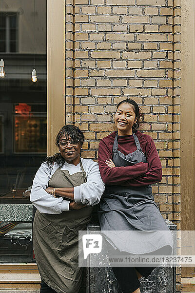 Portrait of smiling female cafe owners wearing aprons standing with arms crossed outside coffee shop