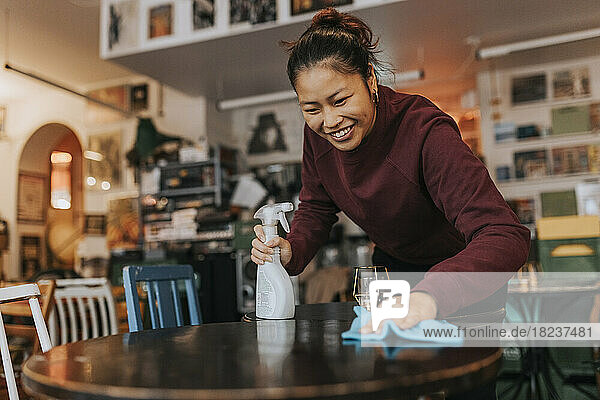 Glückliche Kaffeehausbesitzerin mit Sprühflasche und Lappen beim Reinigen des Tisches im Café