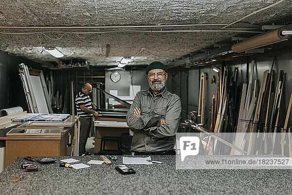 Porträt eines lächelnden männlichen Unternehmers  der mit verschränkten Armen vor einem in einer Werkstatt arbeitenden Kollegen steht