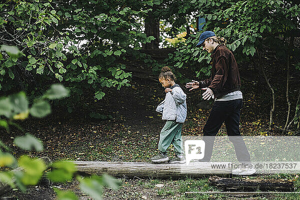 Seitenansicht eines Mädchens  das auf einem Baumstamm balanciert  während ein männlicher Lehrer sie im Park führt
