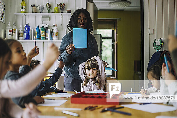 Fröhliche Lehrerin zeigt Schülern farbiges Papier  während sie sie in der Kindertagesstätte unterrichtet
